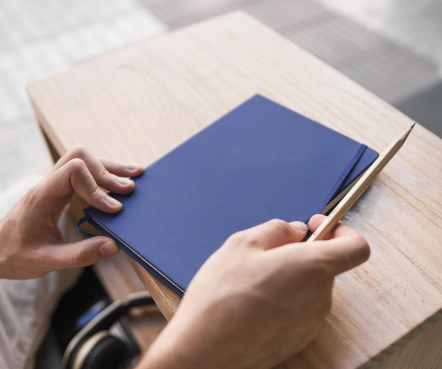Hands holding a blue notebook and pencil at a wooden desk, with headphones nearby — representing personal, hands-on coaching and strategy planning