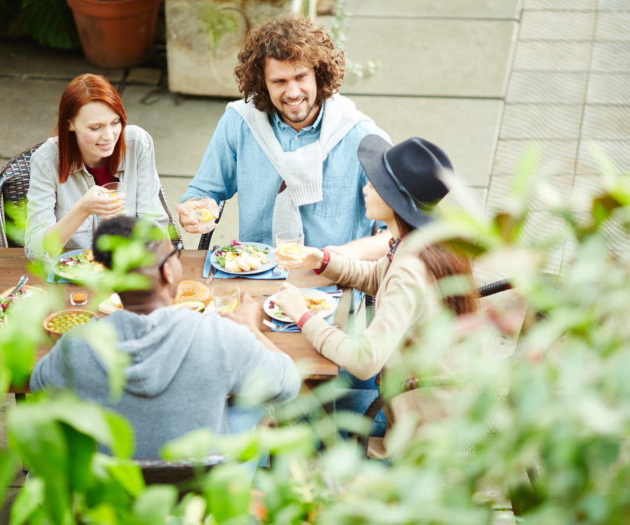 Four people sit around a wooden outdoor table, sharing salads and starters and toasting with glasses of water, surrounded by greenery