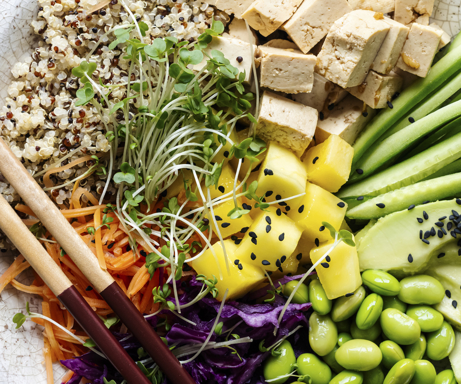 Vegan Buddha bowl from above with quinoa, tofu, mango, cucumber, avocado, edamame, red cabbage, carrots and sprouts, garnished with black sesame, with wooden chopsticks alongside