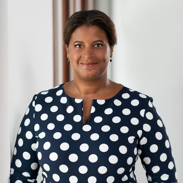 Portrait of Sainey Sawaneh in a navy blue dress with white polka dots, standing indoors and smiling at the camera